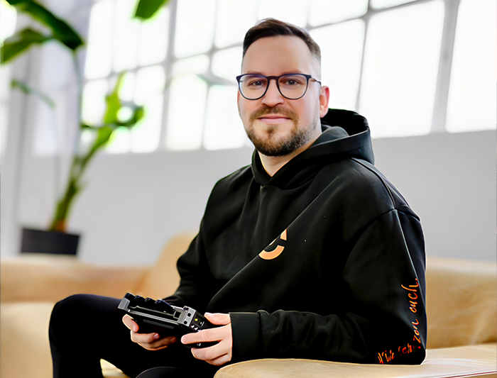 Ein Mann, Florian Böttger, mit Brille sitzt auf einem beigefarbenen Sofa, hält ein elektronisches Gerät in der Hand und trägt einen schwarzen Hoodie mit orangefarbenem Logo von B.CONNECT