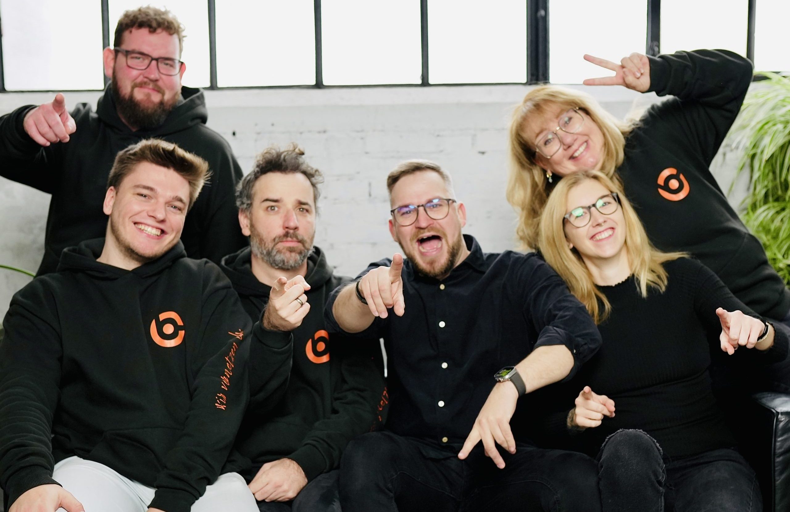 Fröhliche Menschen posieren lachend und gestikulierend vor einem hellen Fenster, teils mit Peace-Zeichen oder Zeigefinger. Mehrere tragen schwarze Hoodies mit orangefarbenem Logo von B.CONNECT